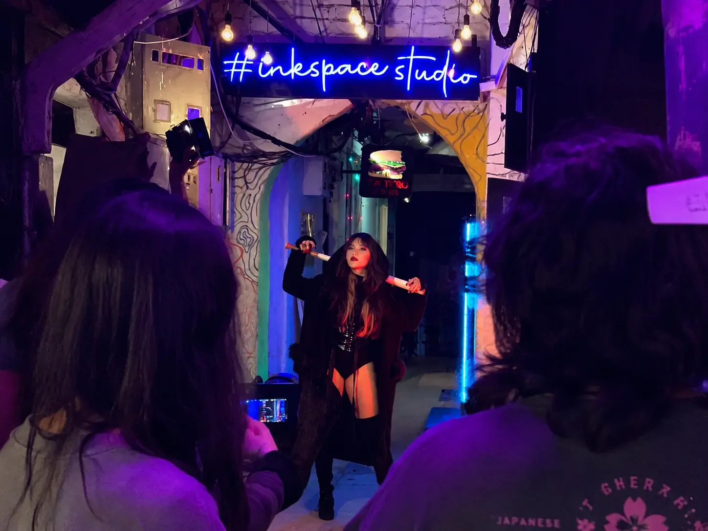 A performer posing under the blue neon #inkspacestudio sign in Space Gully while onlookers film on their phones.