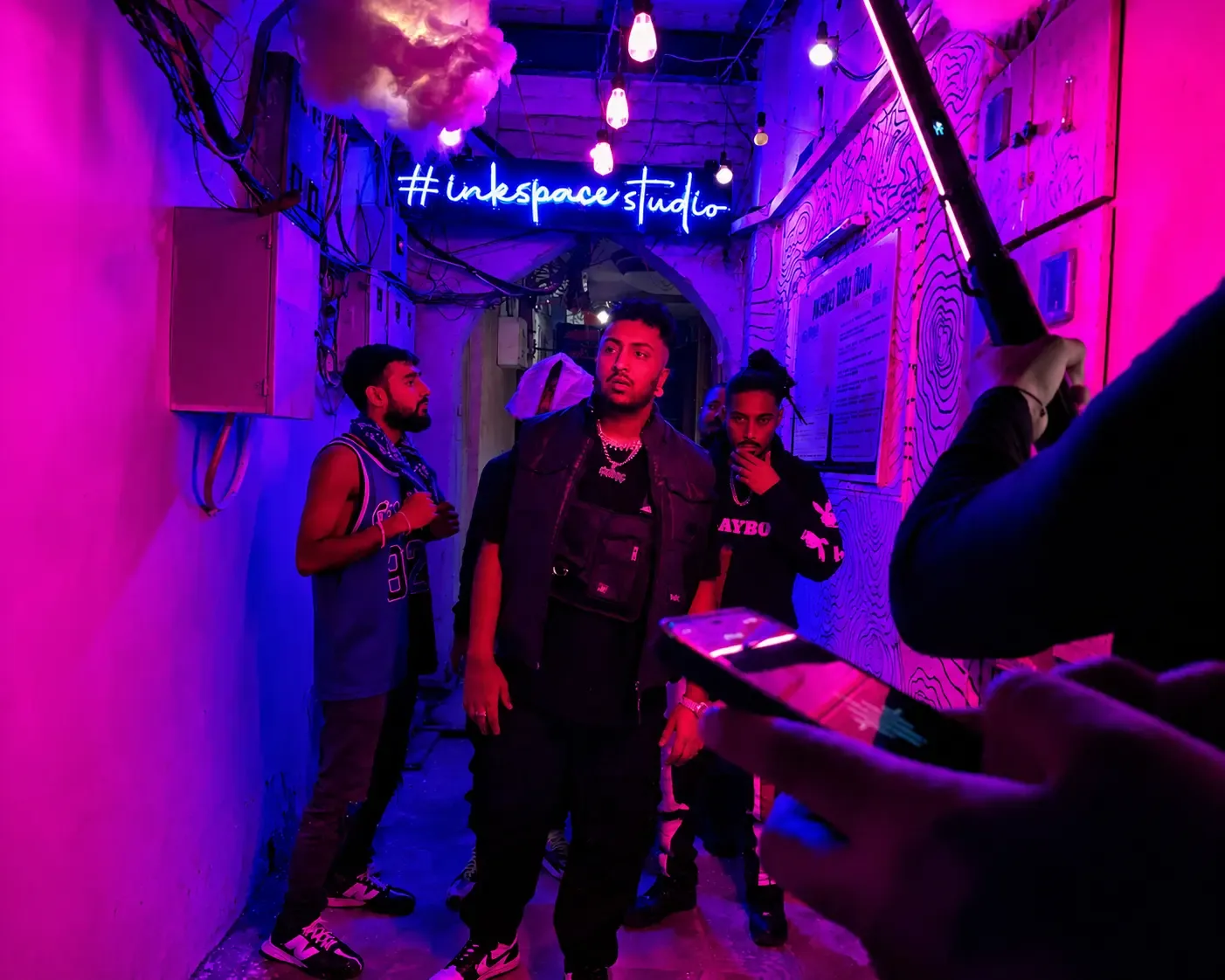 A group of musicians standing in the pink and blue neon-lit Space Gully lane under the #inkspacestudio sign during a video shoot.