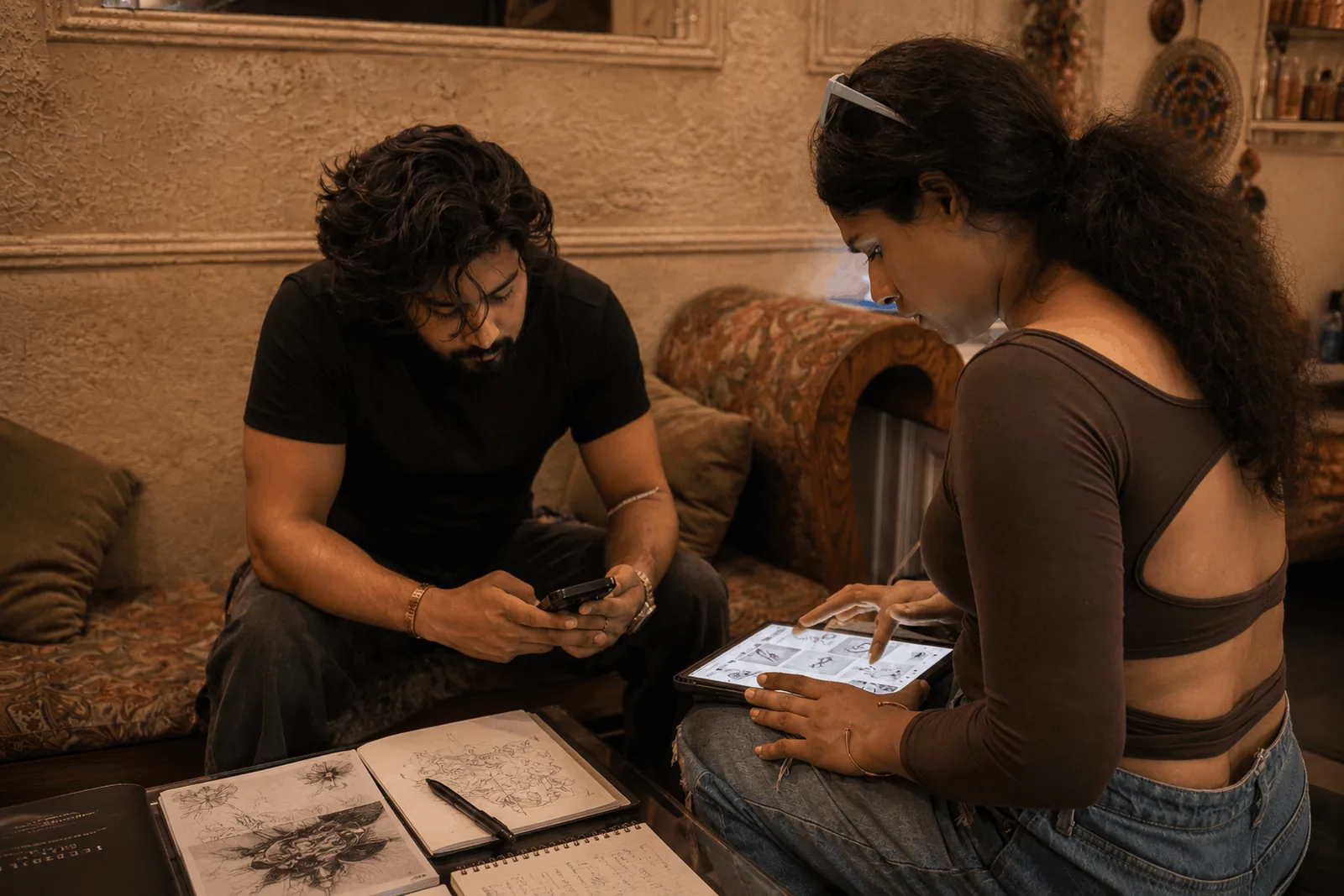 A tattoo artist and a client sitting on a studio sofa, the client scrolling reference sketches on a tablet while open sketchbooks of hand-drawn designs lie on the table in front of them.