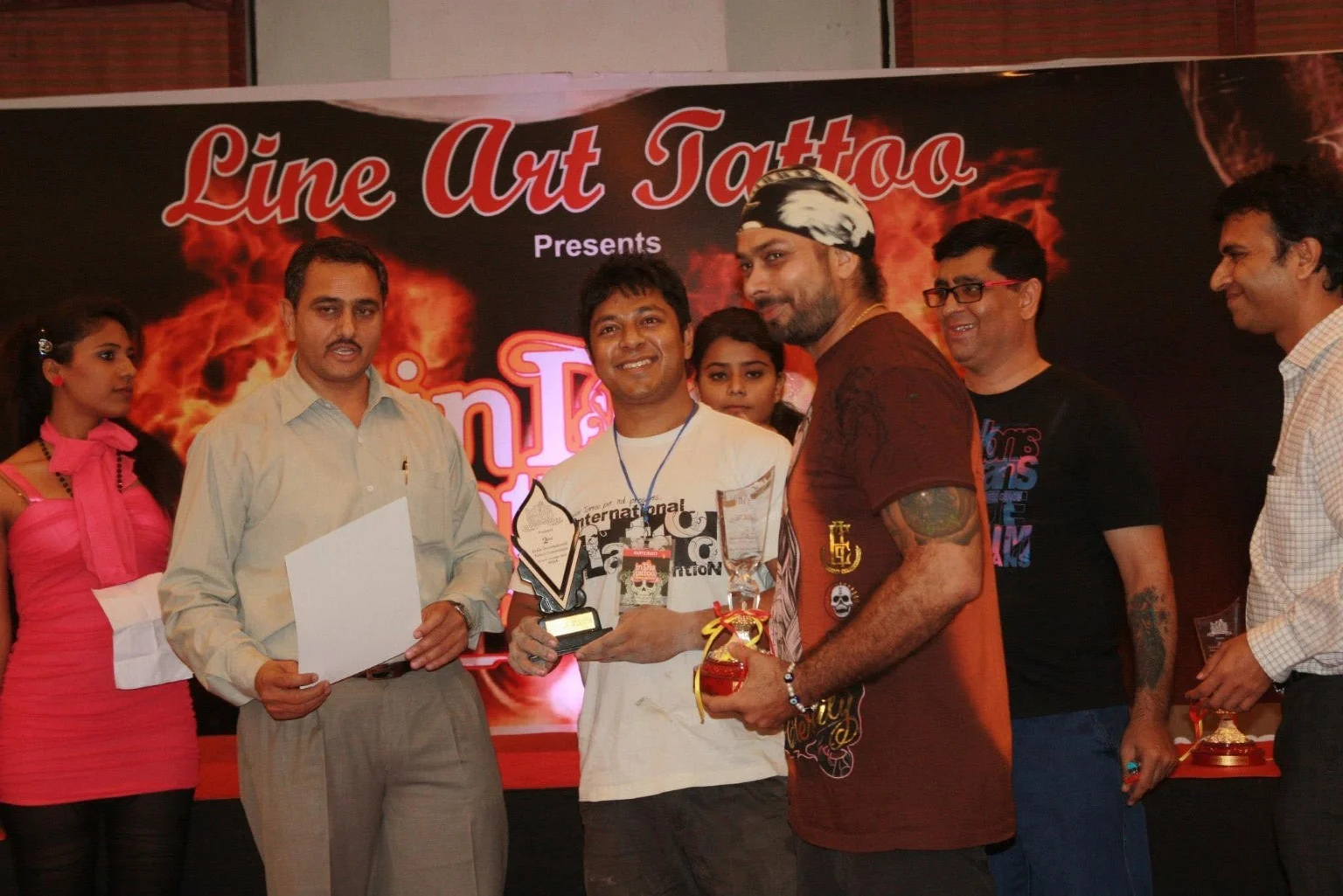 Tattoo artists on stage holding trophies and certificates at a Line Art Tattoo convention award ceremony in India.