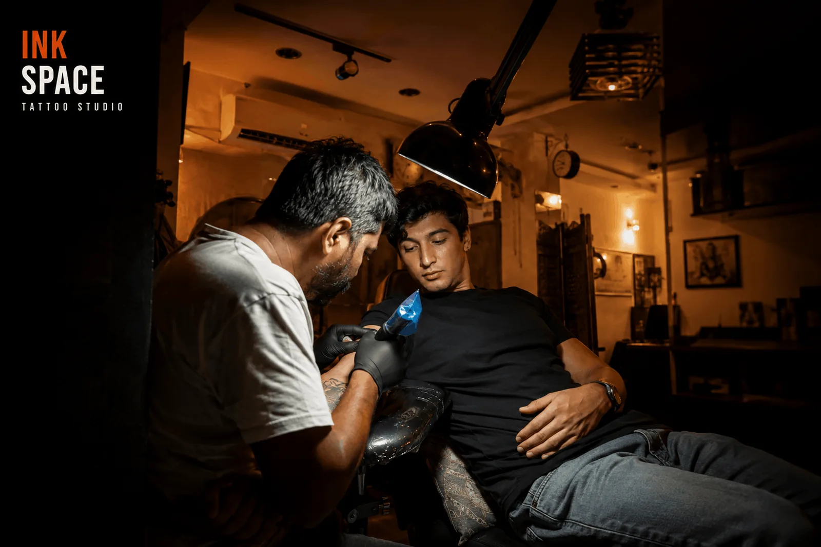 A tattoo artist leaning in with a machine to work on a reclining client's forearm under an angled studio lamp, surrounded by framed artwork and warm studio lighting.