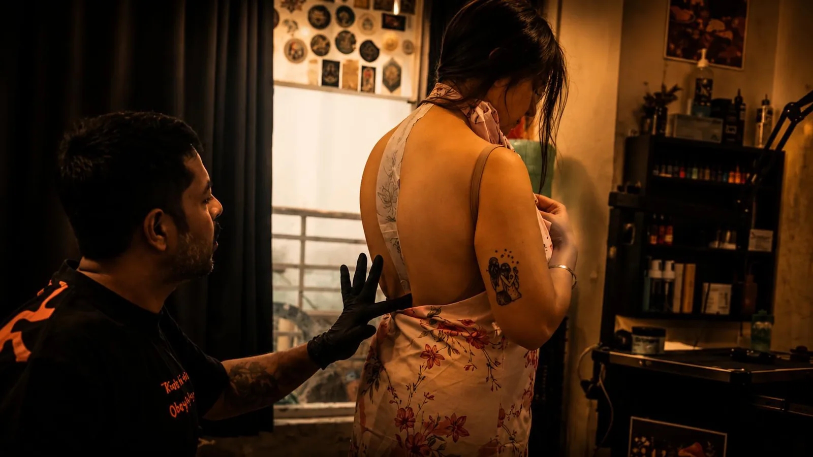 A gloved tattoo artist gesturing towards a standing client's upper back as they discuss where a new piece will sit, with framed tattoo artwork on the studio walls behind them.